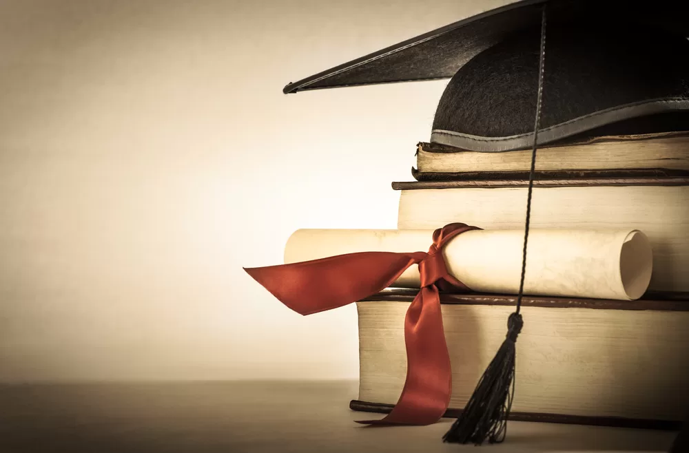 A mortarboard and graduation scroll, tied with red ribbon, on a stack of old battered book