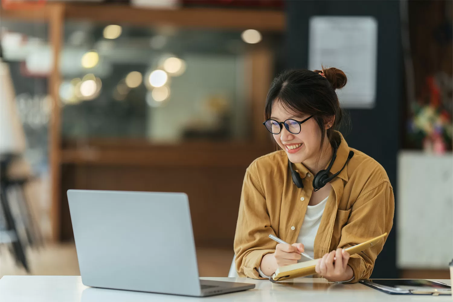 A woman wearing a brown jacket with glasses and headphones looking at a laptop with a pen and a book in hand.